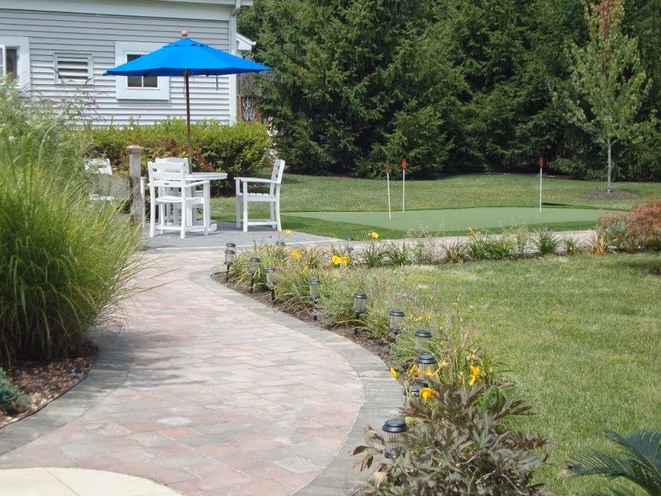 A paved garden pathway curves through a landscaped outdoor area with green grass, flowering plants, and small solar lights. In the background, there is a small putting green with three golf holes and red flags. To the left, there is a patio area with white chairs and a table under a blue umbrella, next to a building with white siding and windows. Tall evergreen trees form a natural backdrop.