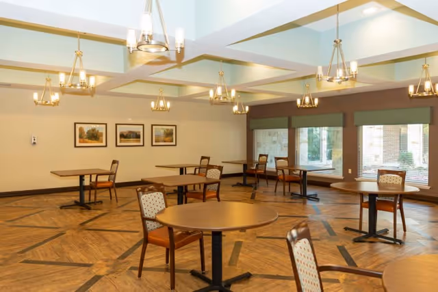 A spacious dining room with multiple round and rectangular wooden tables, each accompanied by chairs with patterned upholstery. The room features large windows with green valances, framed landscape paintings on the wall, and several modern chandeliers hanging from a coffered ceiling. The floor has a decorative wood pattern.