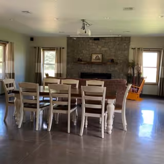 A spacious room with a long wooden dining table surrounded by eight chairs in the foreground. Behind the table is a seating area with a brown couch facing a stone fireplace. The room has large windows with curtains allowing natural light to enter. A rocking chair is placed near one of the windows on the right side. The floor is polished concrete, and a ceiling-mounted projector is visible above the dining table.