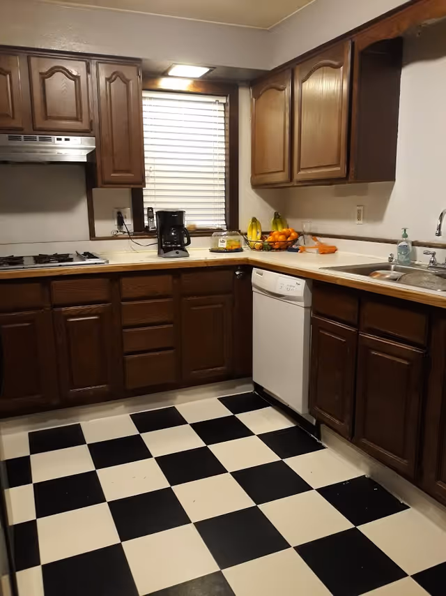 A kitchen with dark wooden cabinets, a black and white checkered floor, a white dishwasher, a sink with a soap dispenser, a coffee maker on the counter, a bowl of fruit, and a window with closed blinds.