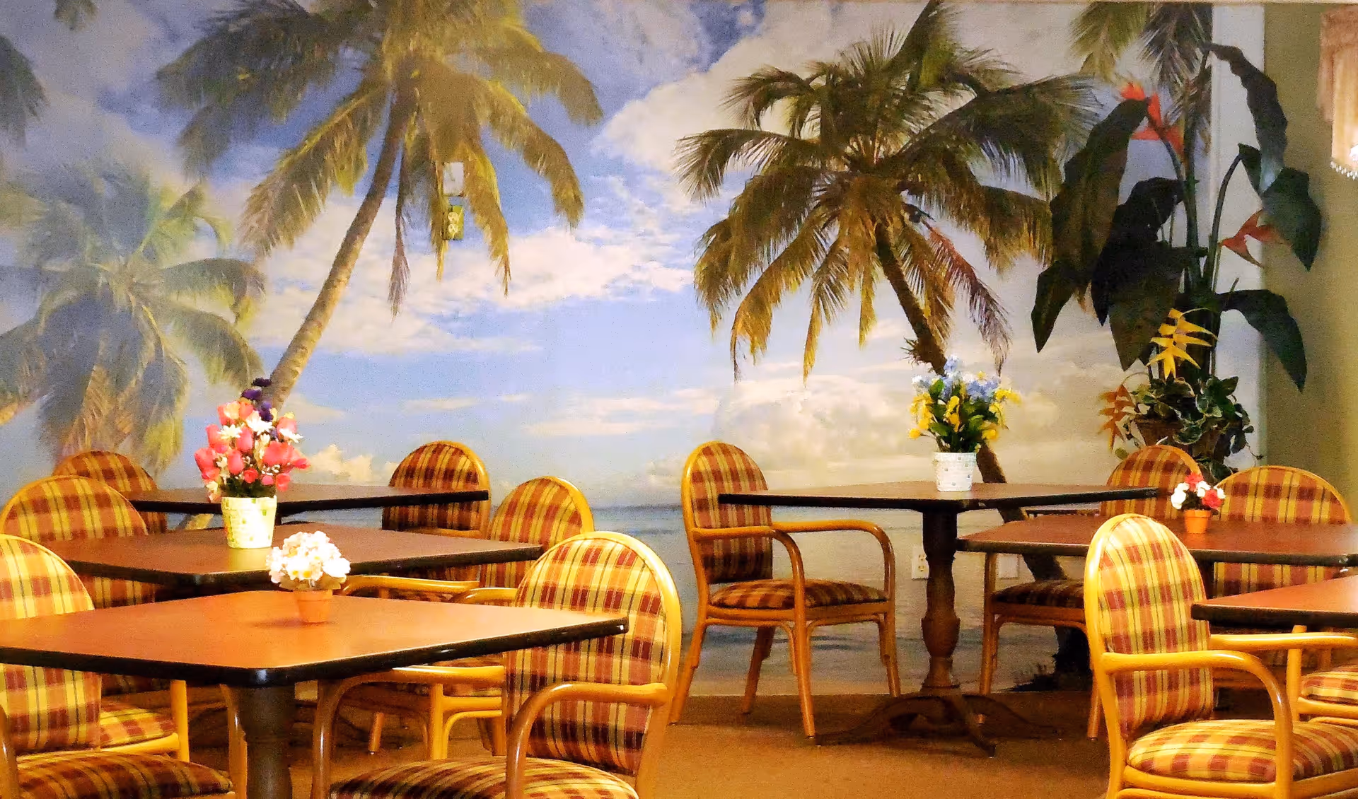 Indoor dining area with several tables and chairs featuring yellow and red plaid upholstery. Each table has a small flower arrangement. The back wall has a large mural of palm trees and a blue sky with clouds, creating a tropical ambiance.