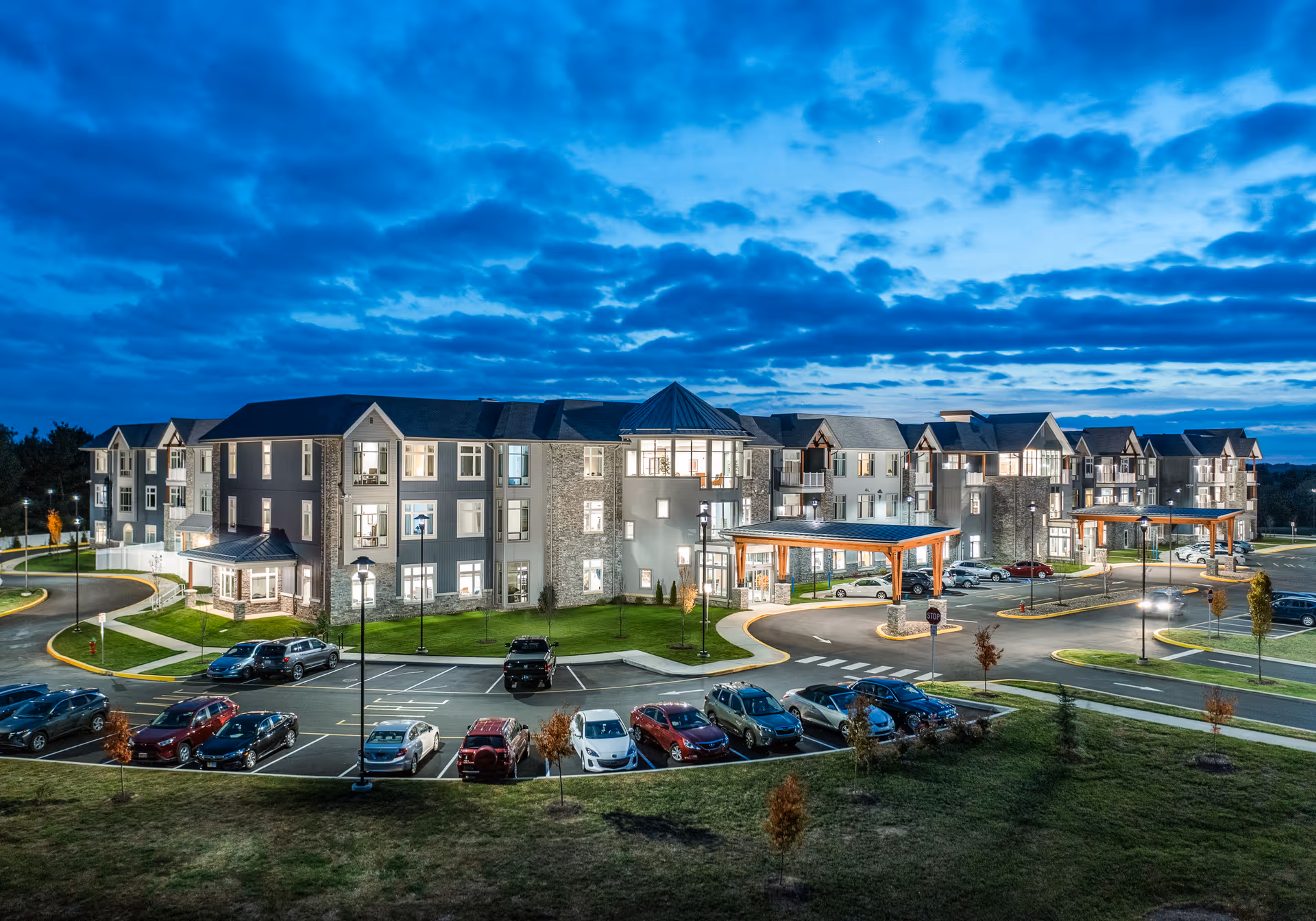 Exterior view of a large, modern senior living facility named Meadowcrest at Middletown during dusk, with well-lit windows, multiple cars parked in the parking lot, and a partly cloudy blue sky overhead.