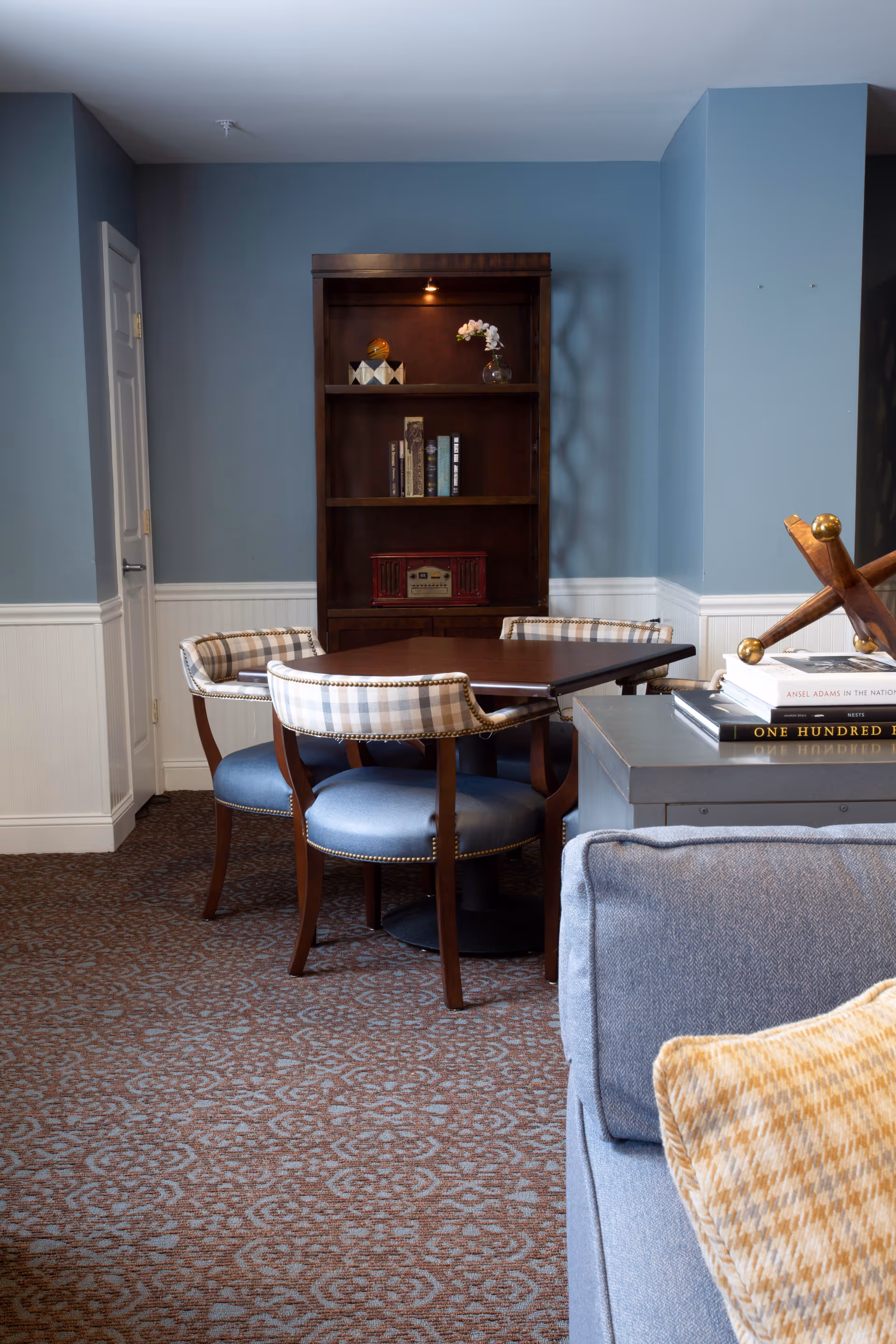 A cozy interior room with a round wooden table surrounded by four upholstered chairs with plaid backs and blue seats. Behind the table is a dark wooden bookshelf with books, a decorative item, and a small vase with flowers. The walls are painted blue with white wainscoting, and a patterned carpet covers the floor. Part of a blue sofa with a yellow patterned pillow is visible in the foreground.