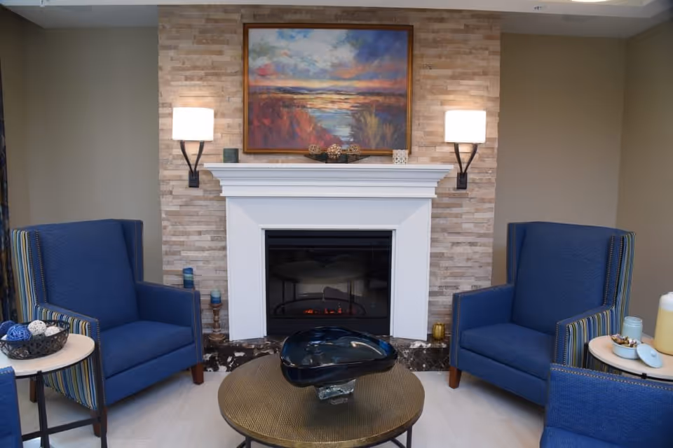 A cozy sitting area with two blue armchairs facing a white fireplace with a stone brick backdrop. Above the fireplace hangs a colorful landscape painting. Two wall-mounted lamps with white shades flank the painting. A round coffee table with a decorative black glass centerpiece is in front of the fireplace. Small side tables next to each armchair hold decorative items.