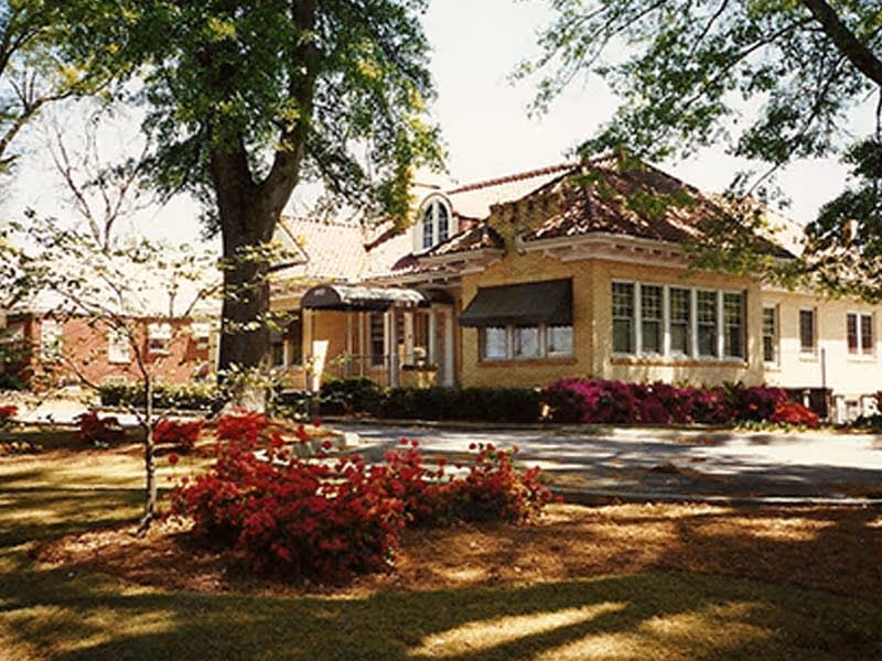 Exterior view of a single-story building with a tiled roof and large windows, surrounded by trees and landscaped garden beds with red flowers, under a sunny sky.