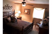 Cozy bedroom with a wooden bed frame, brown bedding, decorative pillows, a ceiling fan with light, a nightstand with a lamp, framed artwork on the walls, a window with curtains, and a wooden dresser with candles and decorative items.