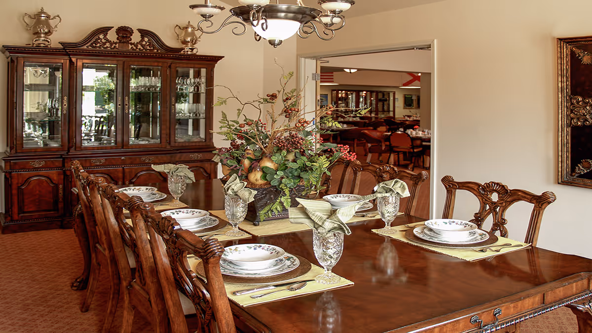 A formal dining room with a polished wooden table set for six people with plates, bowls, crystal glasses, and folded napkins. A large floral centerpiece decorates the middle of the table. In the background, there is a wooden china cabinet filled with glassware and a doorway leading to another dining area with more tables and chairs.