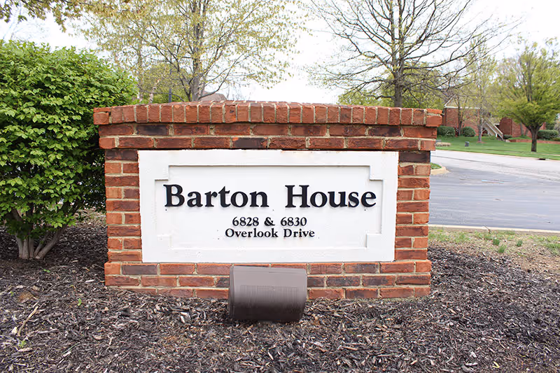 A brick sign with a white inset panel displaying the text 'Barton House 6828 & 6830 Overlook Drive' situated outdoors with some greenery and a paved road in the background.
