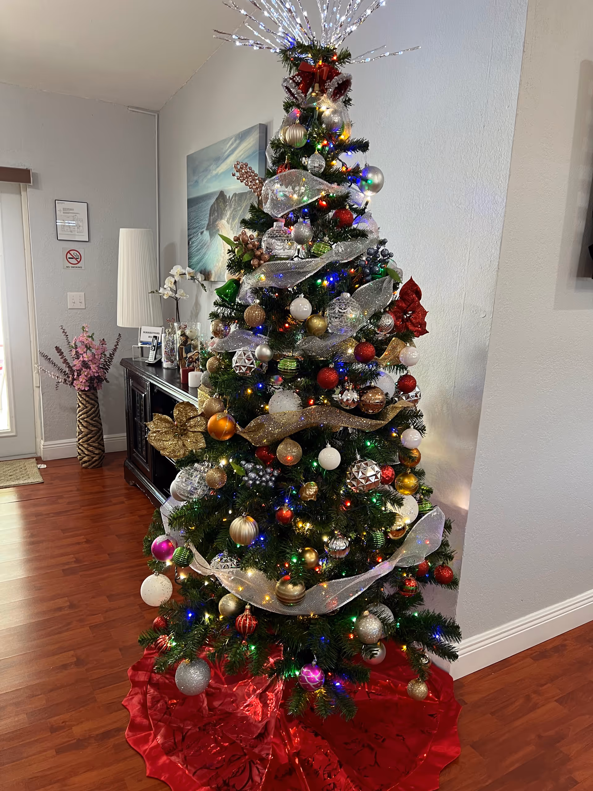 A decorated Christmas tree with multicolored lights, various ornaments, and silver ribbon, standing on a red tree skirt in a room with wooden flooring. In the background, there is a dark wooden cabinet with a white lamp, a framed picture on the wall, and a vase with pink flowers.