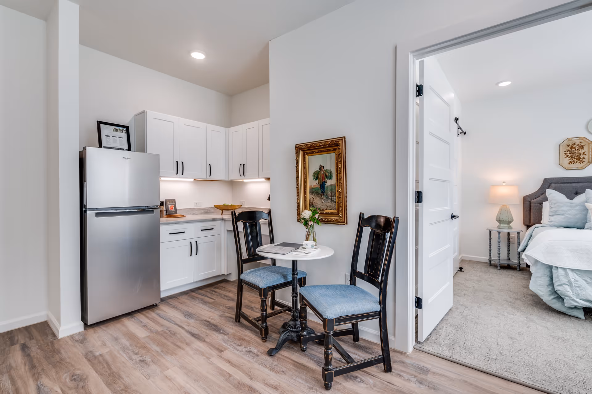 A small kitchen area with white cabinets, a stainless steel refrigerator, and a countertop with under-cabinet lighting. In front of the kitchen is a small round table with two wooden chairs that have blue cushions. A framed painting hangs on the wall above the table. To the right, an open door reveals a bedroom with a bed, bedside table, lamp, and wall decor.