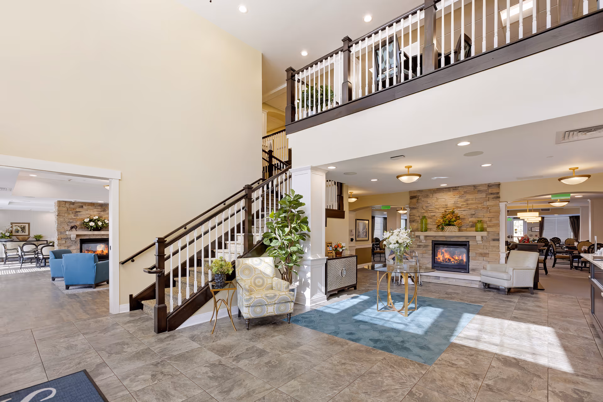 Spacious and well-lit interior of a senior living facility featuring a tiled floor, a staircase with dark wood handrails and white balusters, a stone fireplace with a fire burning, comfortable seating including armchairs, a small table with a flower arrangement, and an open layout leading to dining and lounge areas.