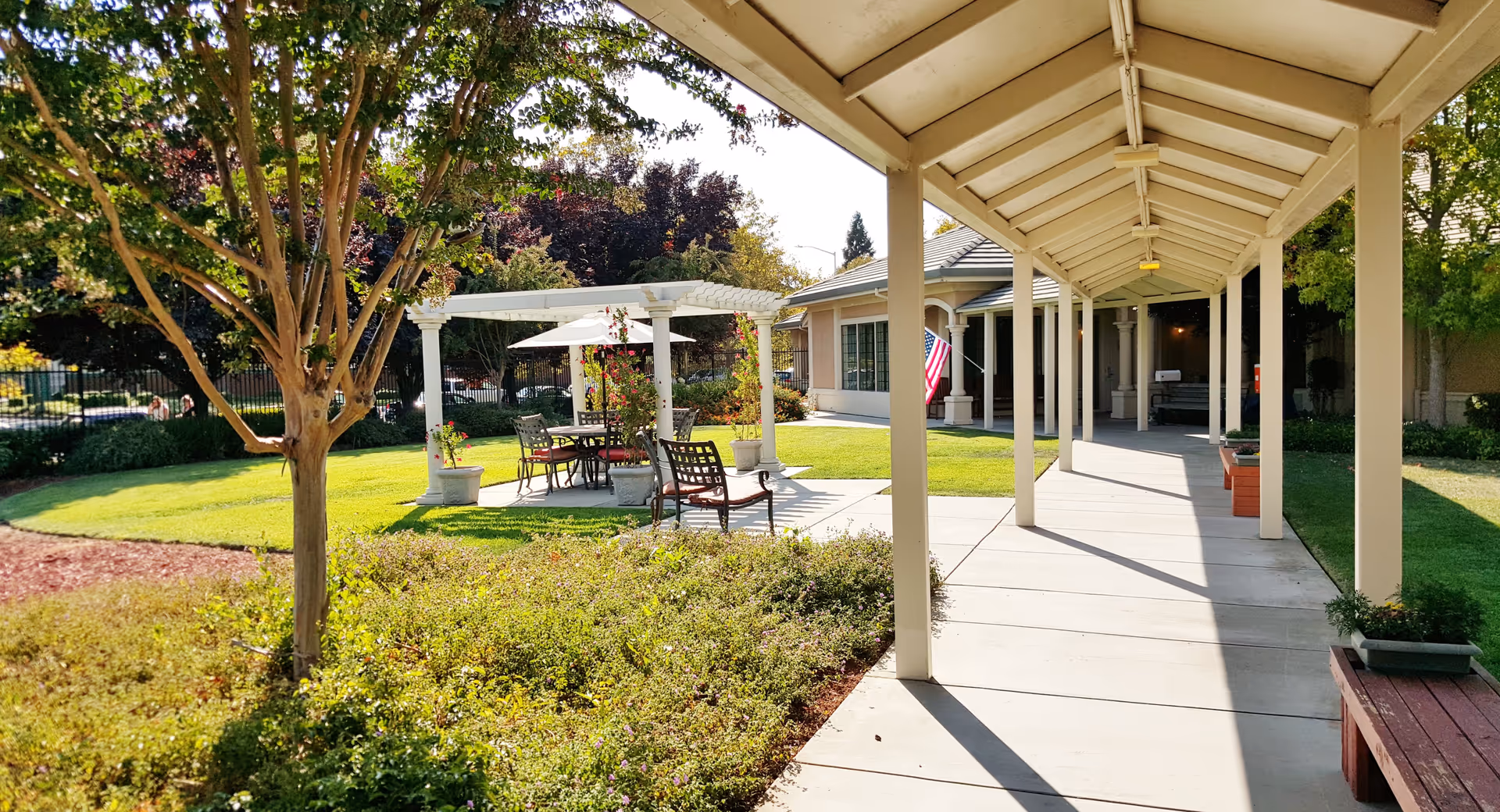 A covered walkway leads through a landscaped courtyard with a pergola seating area, benches, lawn, and an American flag by the building entrance.