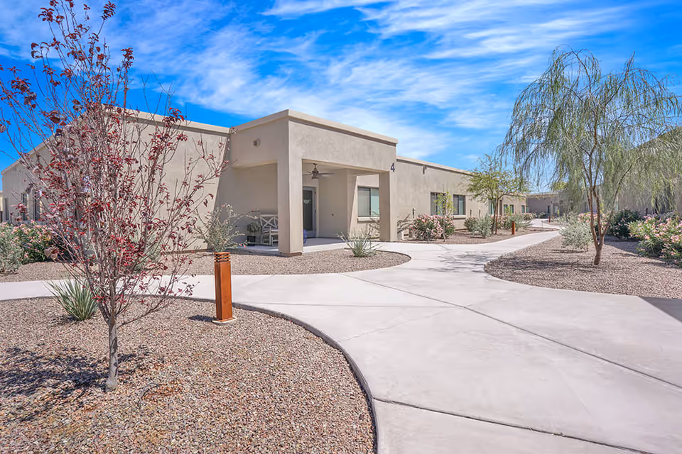Exterior view of a single-story senior living facility building with beige stucco walls, a covered entrance with a ceiling fan, and a concrete walkway surrounded by desert landscaping including small trees and shrubs under a blue sky with scattered clouds.