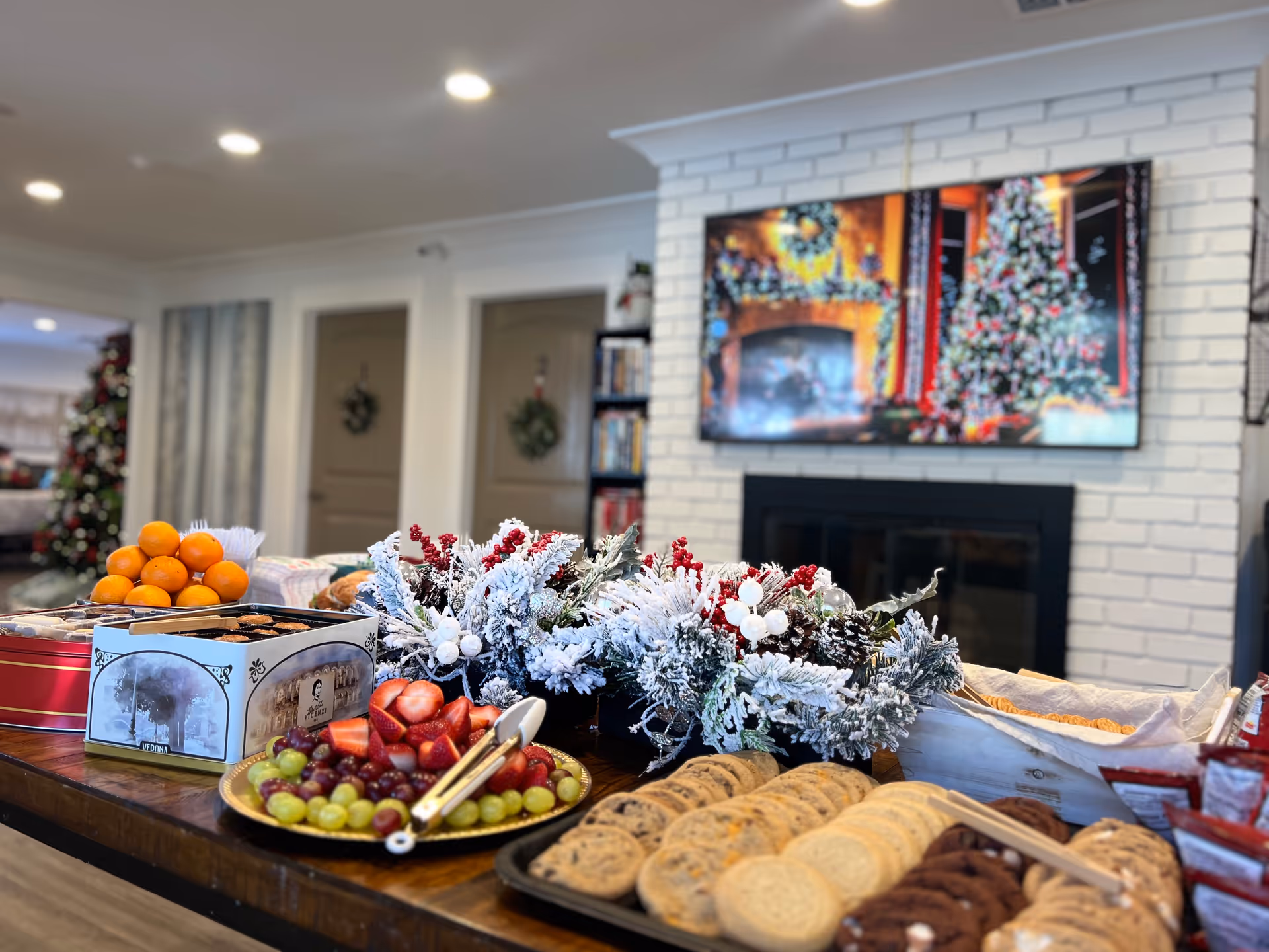 A festive snack table with fruit, cookies and a snowy holiday centerpiece in a senior living common room with a TV above a fireplace.