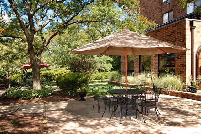 Outdoor patio area at Westwood Place with a round metal table and six chairs under a large beige umbrella. The patio is surrounded by greenery including trees, bushes, and ornamental grasses, with a brick building visible in the background.