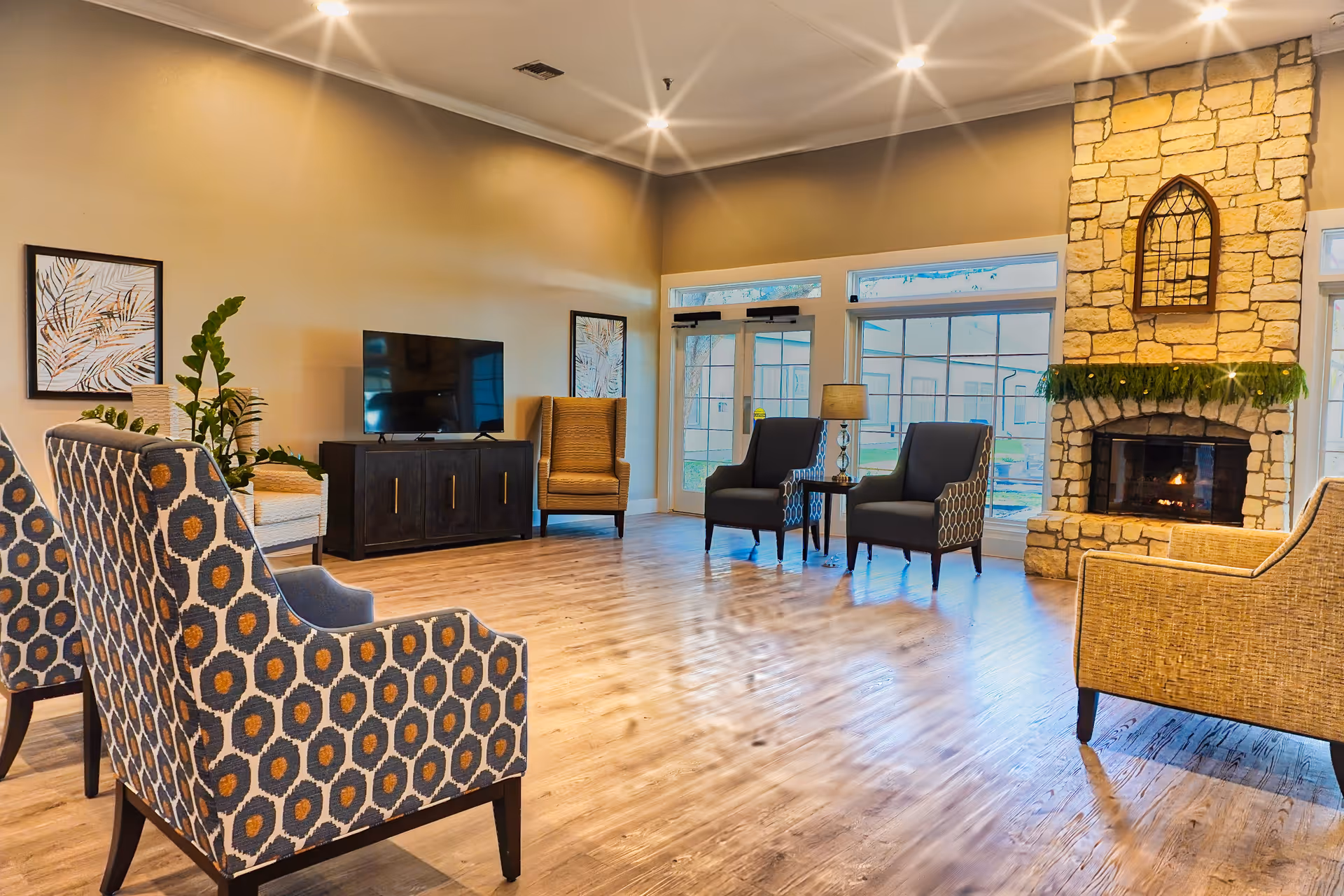 A spacious living room with wooden flooring, patterned armchairs, a stone fireplace with a green garland, a TV on a dark wooden cabinet, and large windows letting in natural light.