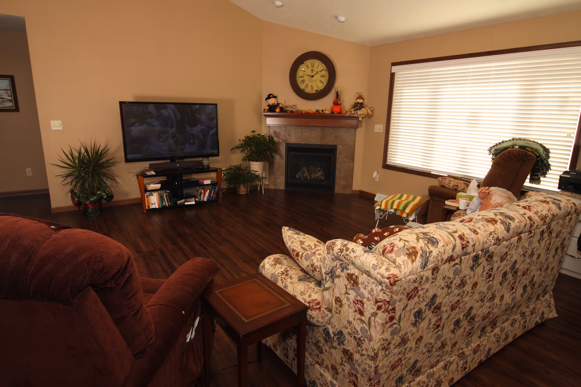 Cozy living room with a floral sofa and recliners facing a TV and fireplace, a large window with blinds, and a person seated in a recliner.
