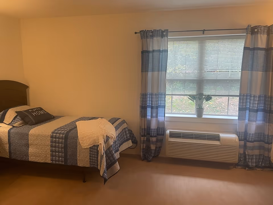 Simple furnished bedroom with a single bed covered in blue patterned bedding, a window with striped curtains and a wall air conditioner below.