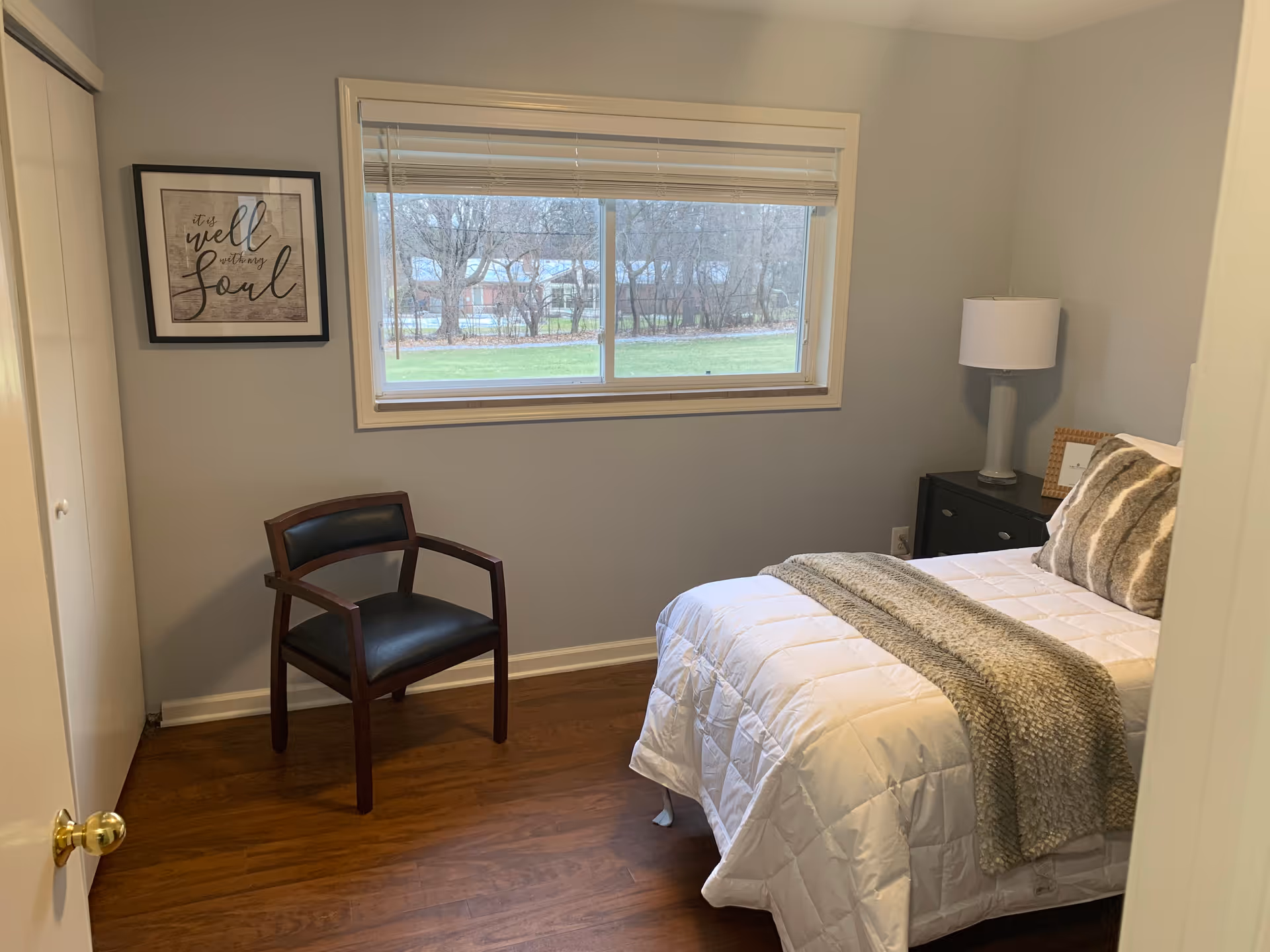 A small bedroom with a single bed covered in a white comforter and a gray throw blanket. There are two gray and white pillows on the bed. Next to the bed is a black nightstand with a white lamp and a small picture frame. A wooden chair with black cushioning is placed near the wall. A window with white blinds shows a view of trees and grass outside. A framed wall art with the text 'It is well with my soul' hangs on the wall.