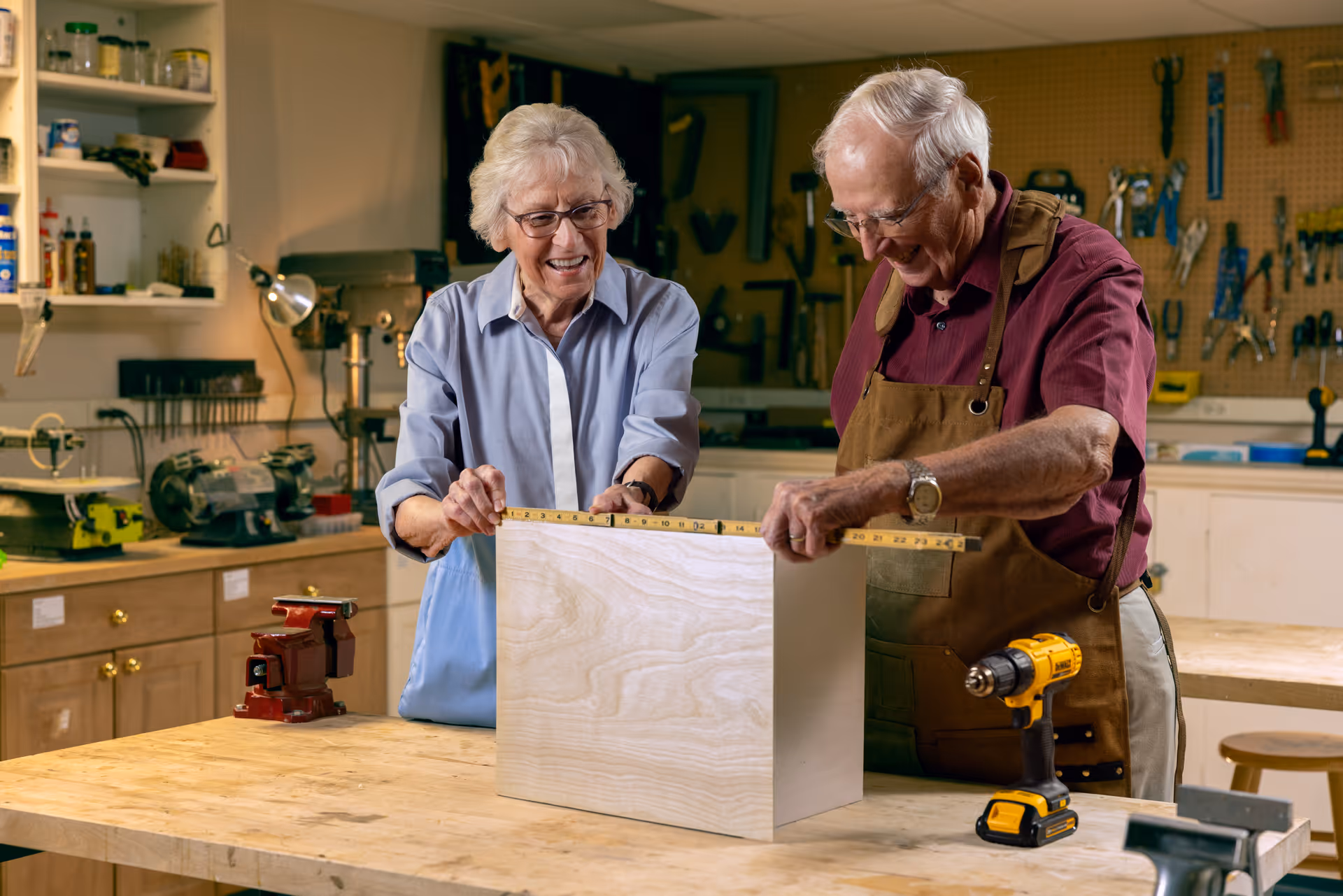 An elderly man and woman in a woodworking workshop measuring a wooden box together. The man is wearing a brown apron and a maroon shirt, while the woman is wearing glasses and a light blue shirt. Various tools and equipment are visible in the background.