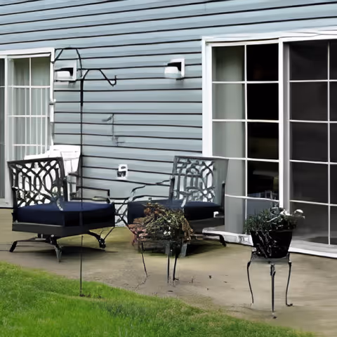 Outdoor patio area with two metal chairs with dark cushions, a small glass-top table, and two plant stands with potted plants. The patio is adjacent to a building with light blue siding and large sliding glass doors.