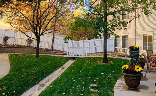 Well-maintained courtyard with green lawn, trees, a white fence, potted flowers, and seating next to a beige building.