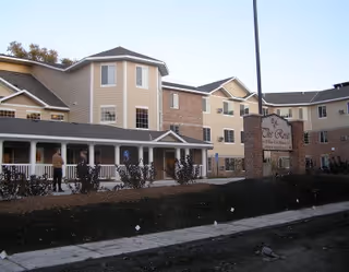 Exterior view of a multi-story senior living facility building with beige and brick facade, a covered porch with white railings, and a sign that reads 'The Rose' near the entrance. There are bushes and a sidewalk in front of the building.