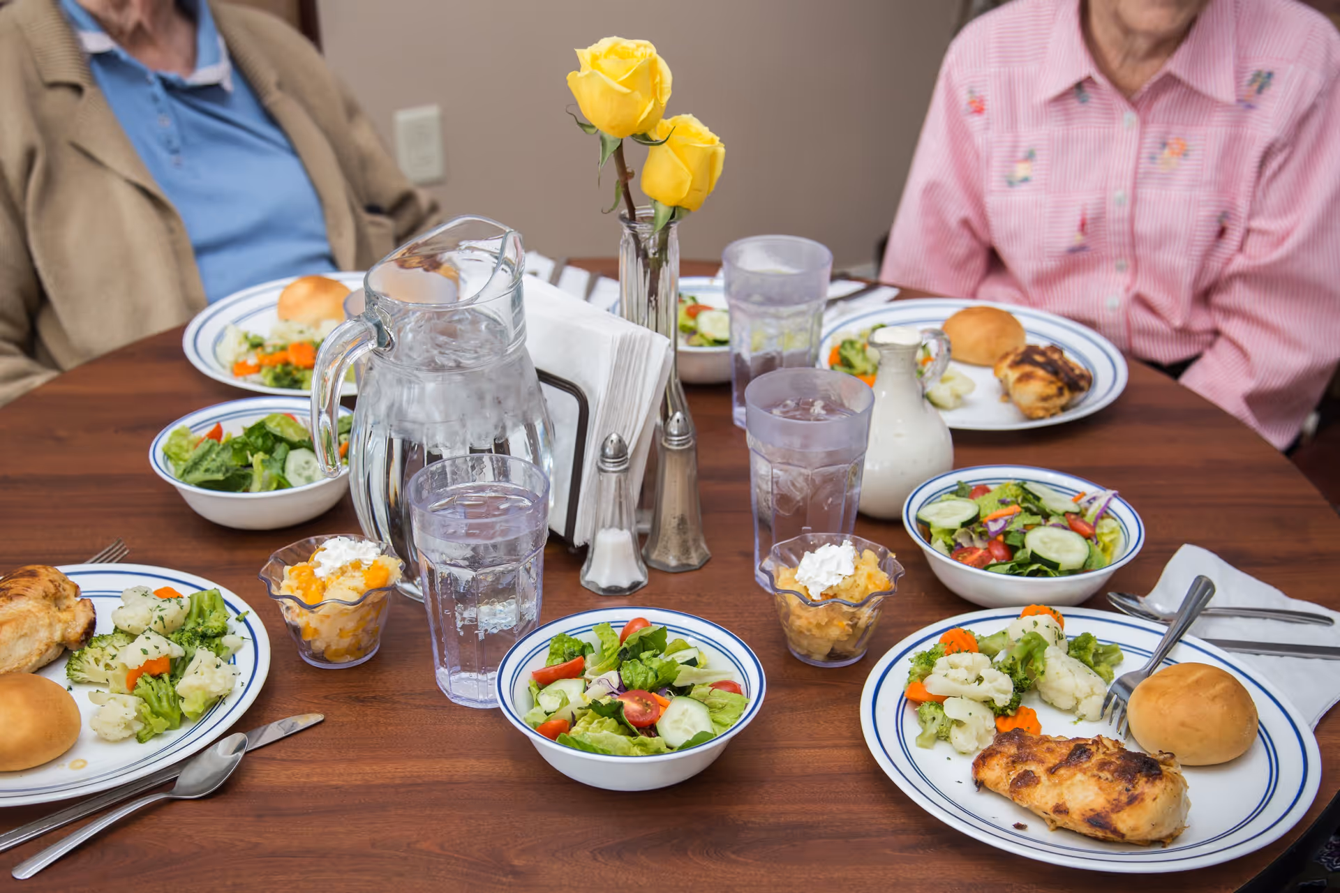 A dining table set with plates of food including grilled chicken, mixed steamed vegetables, salad bowls, bread rolls, and glasses of water. A pitcher of water, a small vase with two yellow roses, salt and pepper shakers, and napkins are also on the table. Two elderly people are partially visible sitting at the table.