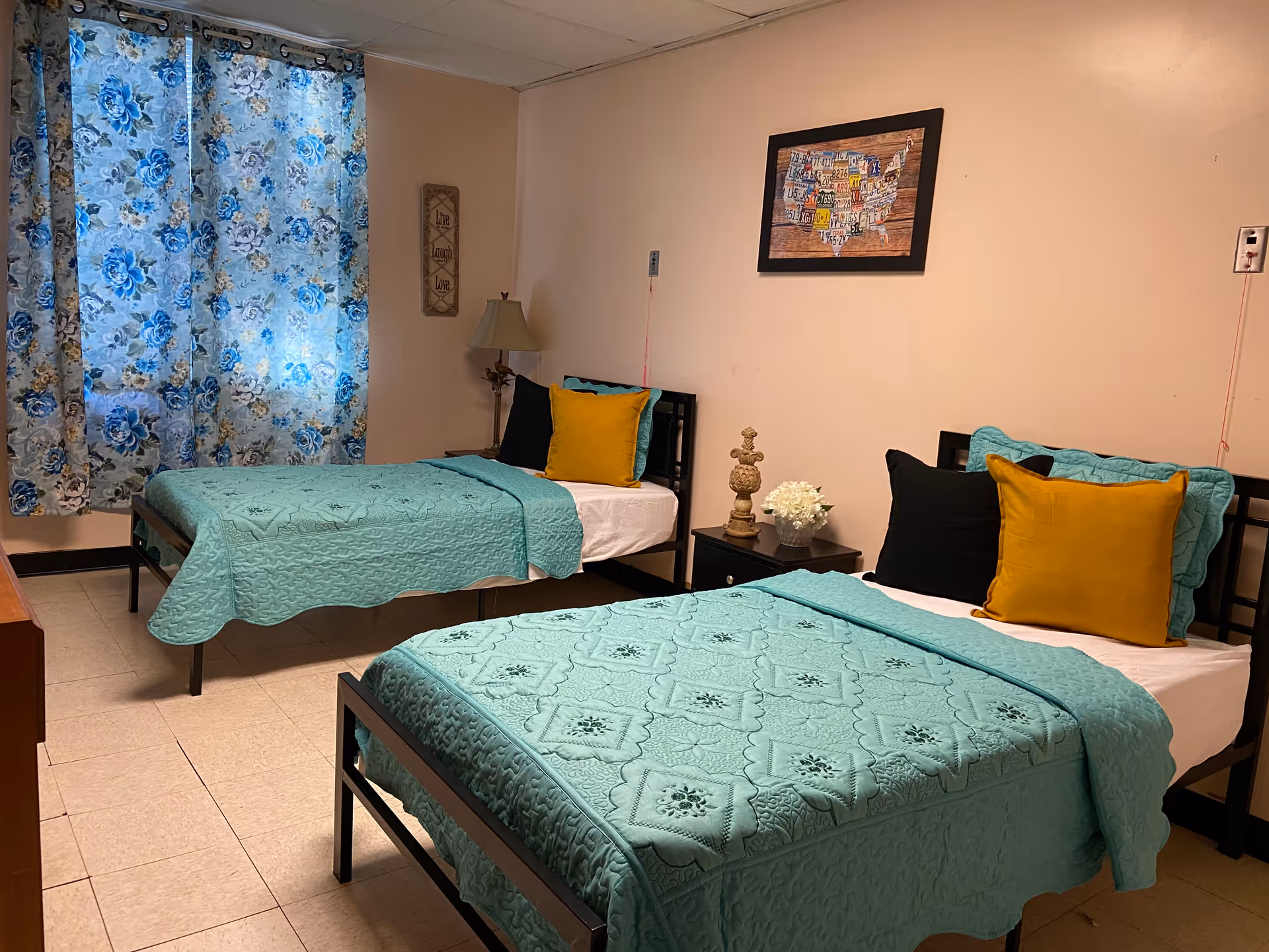 Two twin beds with turquoise quilts, matching pillows, and a floral blue curtain in a bedroom.