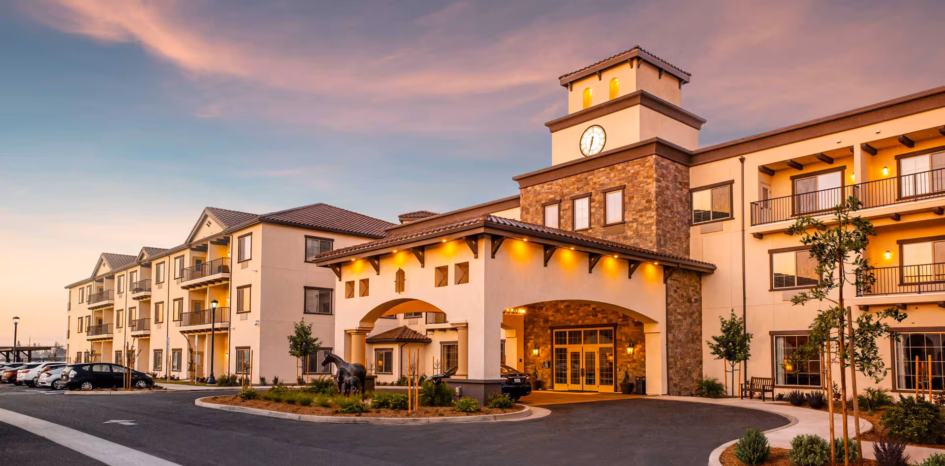 Exterior view of The Park at Modesto Independent Living Community building at sunset, featuring a three-story structure with balconies, a clock tower, and a covered entrance with warm lighting. There are parked cars and landscaped greenery around the driveway.