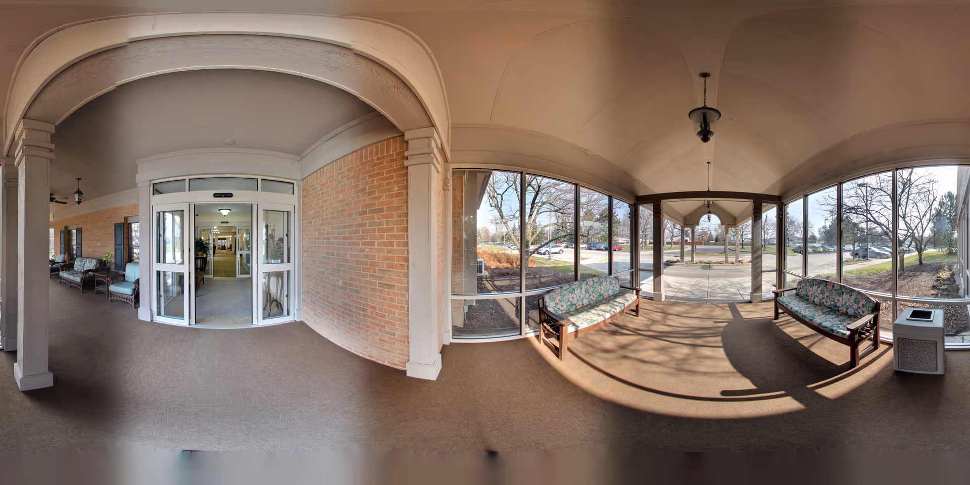 Covered entrance area of Oakmont Livonia Senior Community with glass sliding doors leading inside. The area has cushioned benches with floral patterns on both sides and large windows providing a view of the outdoor parking lot and trees.