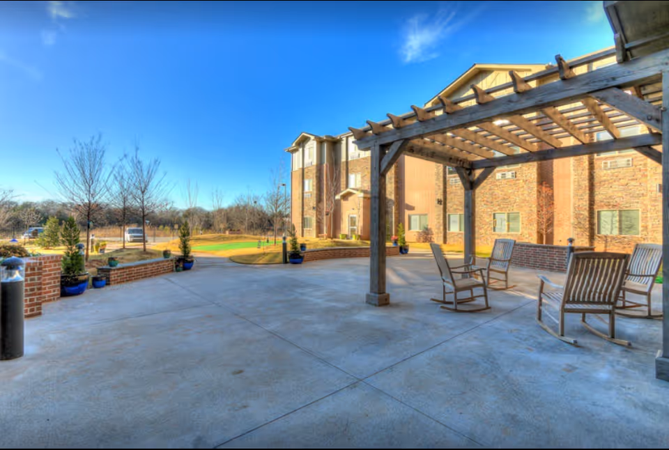 Outdoor patio with a wooden pergola and rocking chairs in front of a brick senior living building under a blue sky.