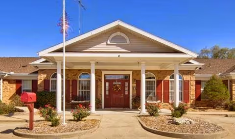 Front exterior view of a single-story building with a peaked roof, white columns supporting a covered entrance, red double doors decorated with a wreath, red shutters on windows, a red mailbox, and an American flag on a flagpole in front.