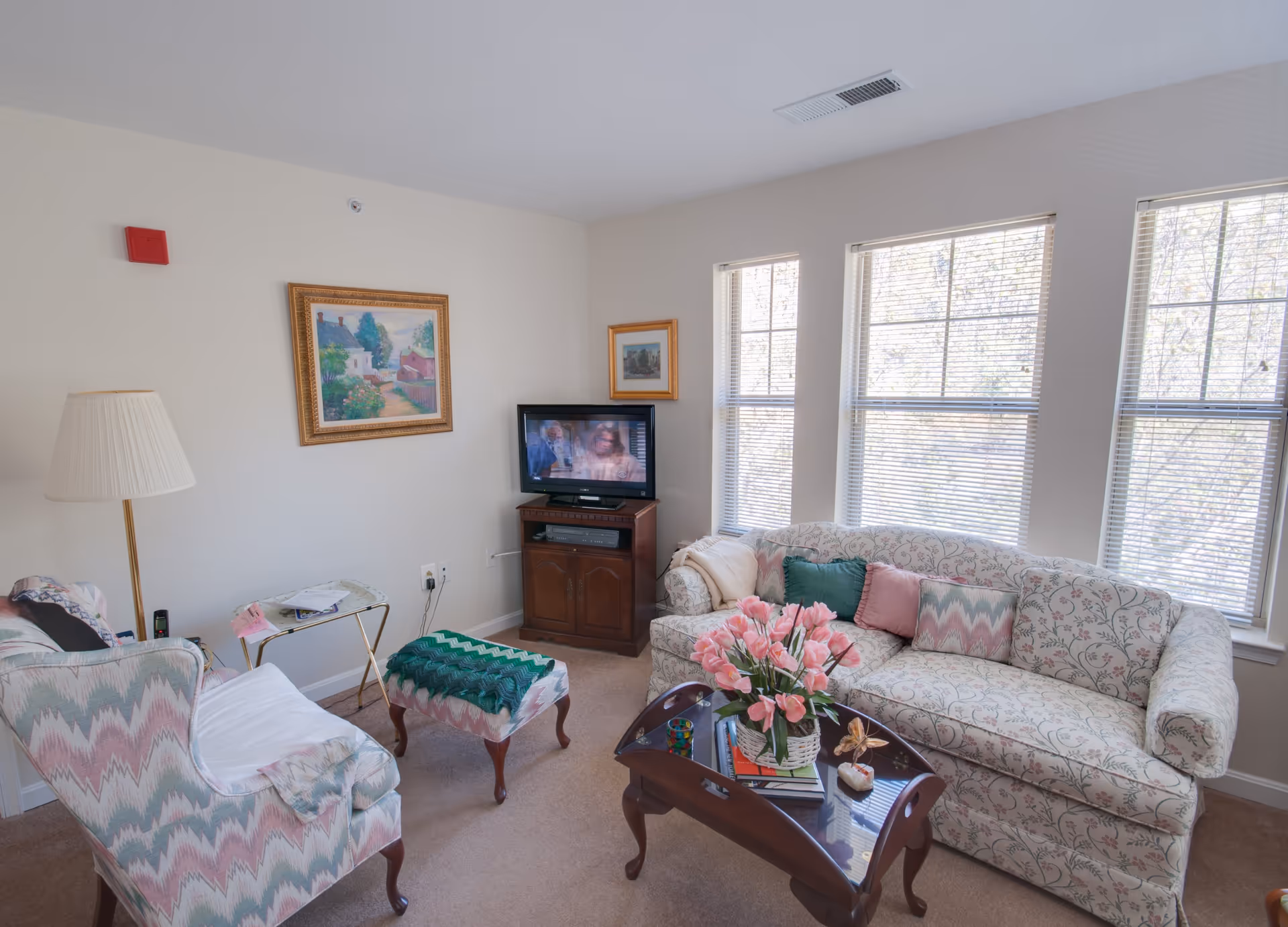 Bright living room with a floral sofa and armchair, coffee table with a vase of pink flowers, a TV on a stand, and three tall windows letting in light.