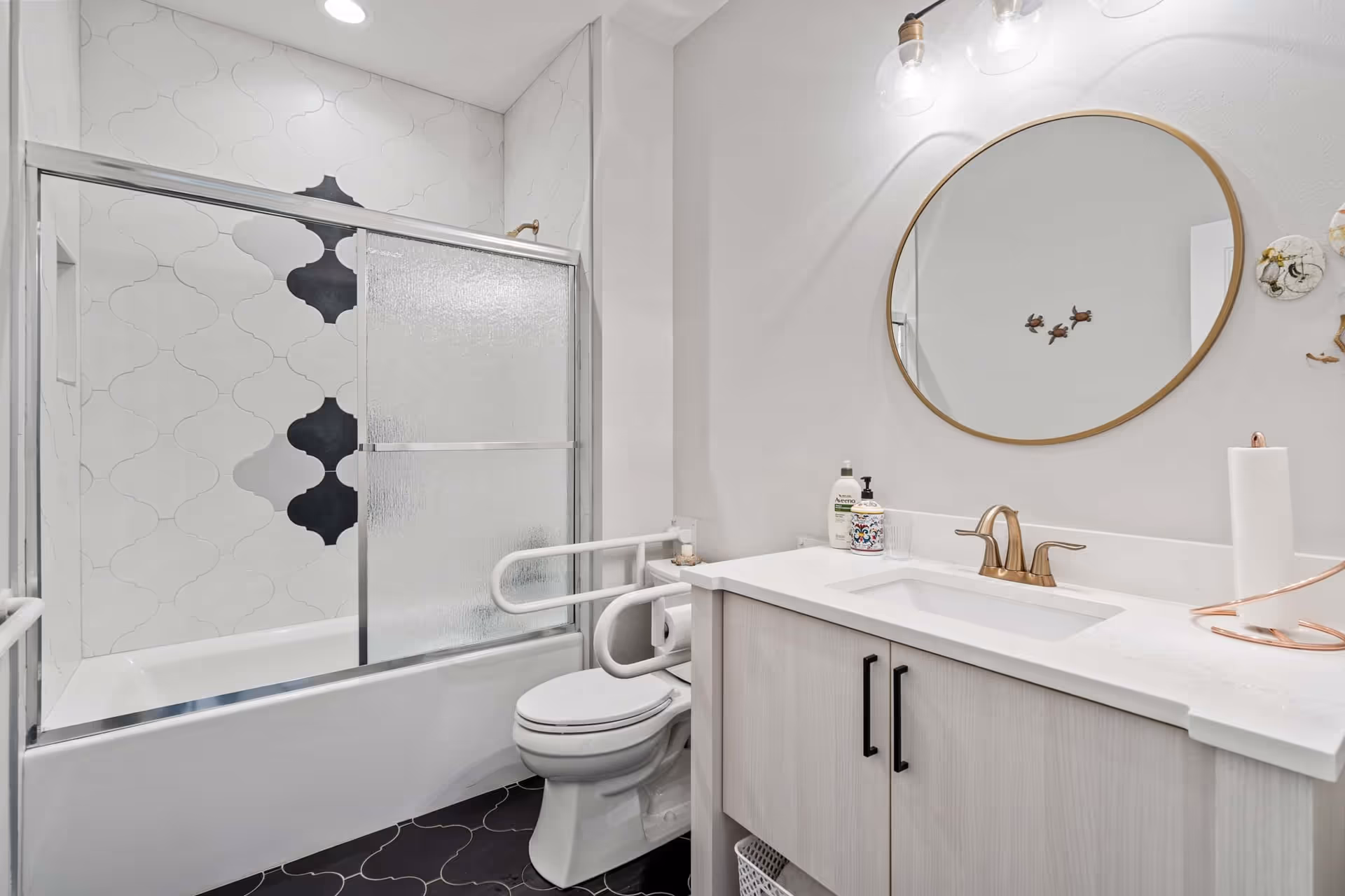 A modern bathroom featuring a bathtub with sliding frosted glass doors and decorative white and black tiles on the wall. There is a white toilet with safety grab bars installed on both sides. A vanity with a white countertop, a gold faucet, and a round mirror with a gold frame is visible. The floor has dark tiles with a light pattern, and there are toiletries and a paper towel holder on the countertop.