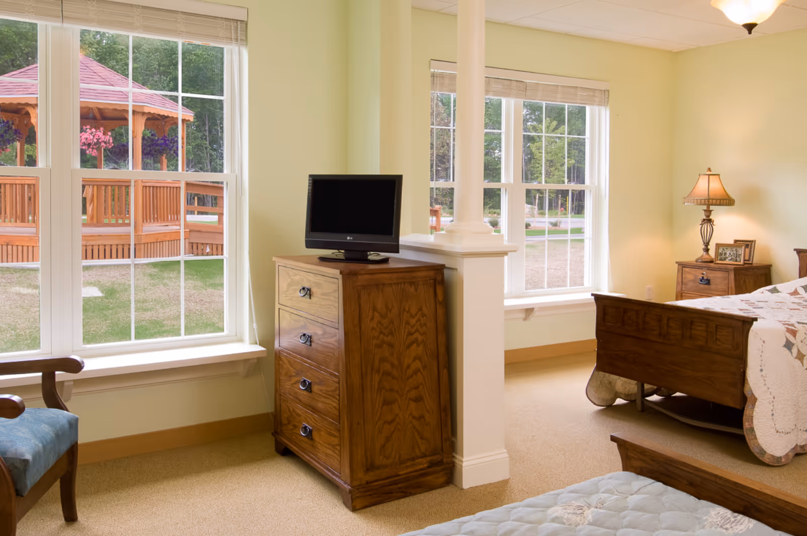 A bright bedroom with large windows showing a wooden gazebo outside. The room features a wooden bed with a quilt, a wooden dresser with a small TV on top, a wooden nightstand with a lamp and framed photo, and a blue cushioned chair.