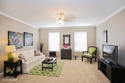 A bright living room with beige walls and carpet, featuring a white sofa with floral pillows, a green patterned rug, a black coffee table, a black side table with a green lamp, two floral paintings above the sofa, a black dresser with a decorative mirror and pink flowers between two windows, a green upholstered chair, and a flat-screen TV on a black TV stand.