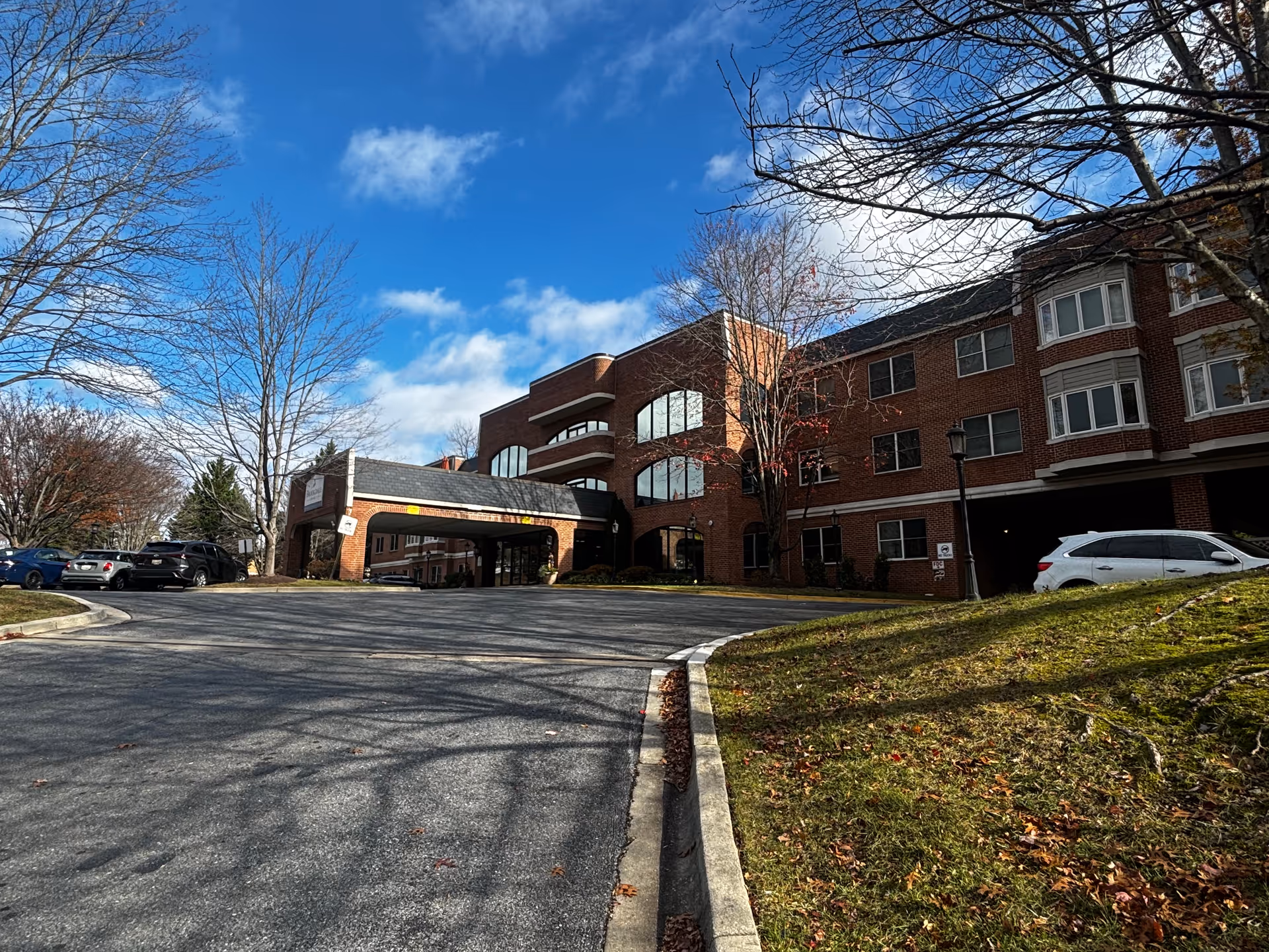Brick multi-story senior living building with a covered drive-through entrance, parked cars, leafless trees, and a blue sky.