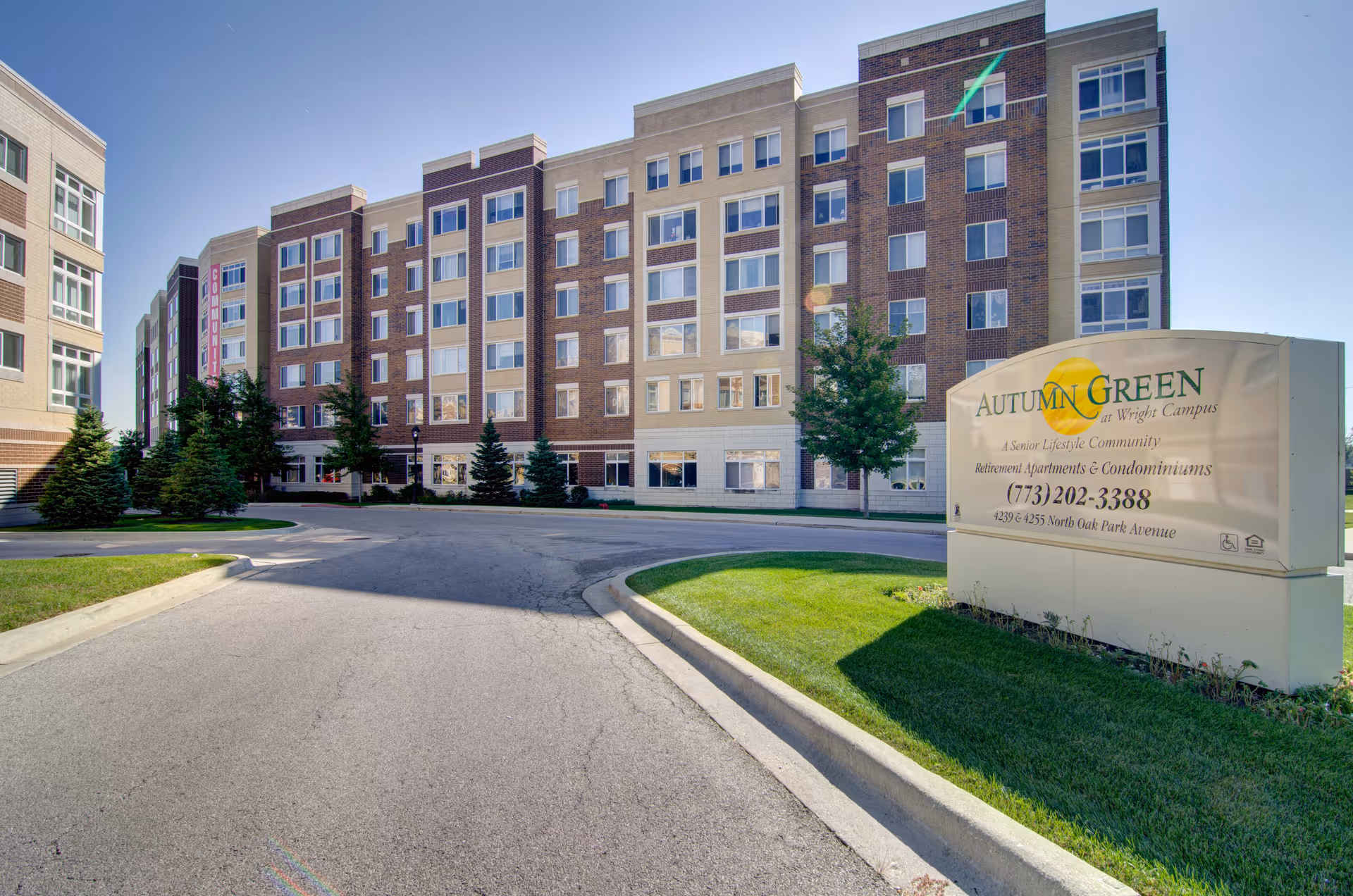 Exterior view of a multi-story senior living facility building with a sign in the foreground that reads Autumn Green at Wright Campus, a senior lifestyle community offering retirement apartments and condominiums. The building is surrounded by a paved driveway, green grass, and trees under a clear blue sky.