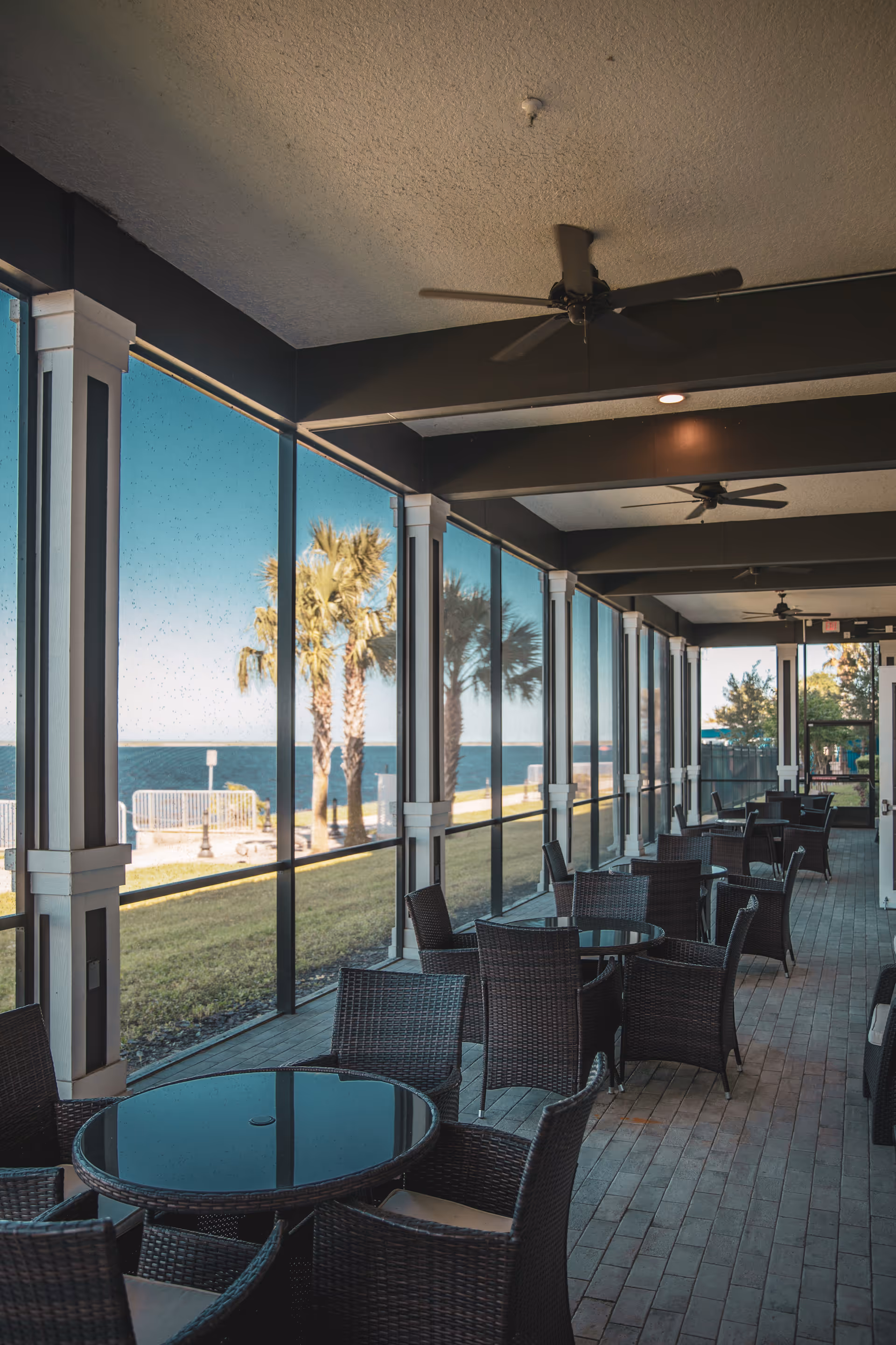 A covered outdoor seating area with multiple round glass tables and wicker chairs. The area is enclosed with large glass panels offering a view of palm trees, grass, and a body of water in the background. Ceiling fans are mounted on the ceiling.