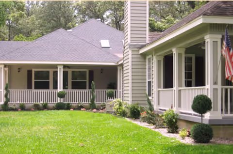 Exterior view of a senior living facility showing a well-maintained lawn, a porch with white railings, and an American flag mounted on the corner of the building. The building has a gray roof and beige siding with trees in the background.