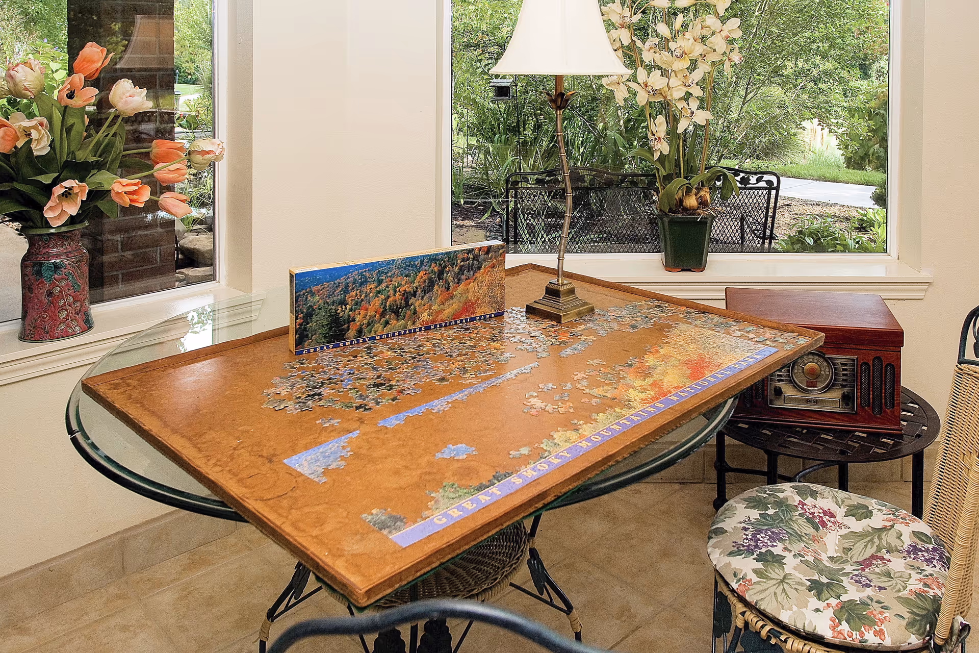 A cozy indoor seating area with a glass-top round table holding a partially completed jigsaw puzzle of a scenic landscape. The table is surrounded by chairs with floral cushions. A vintage radio sits on a small side table next to the main table. There are two large windows showing greenery outside, and flower arrangements are placed on the windowsills.