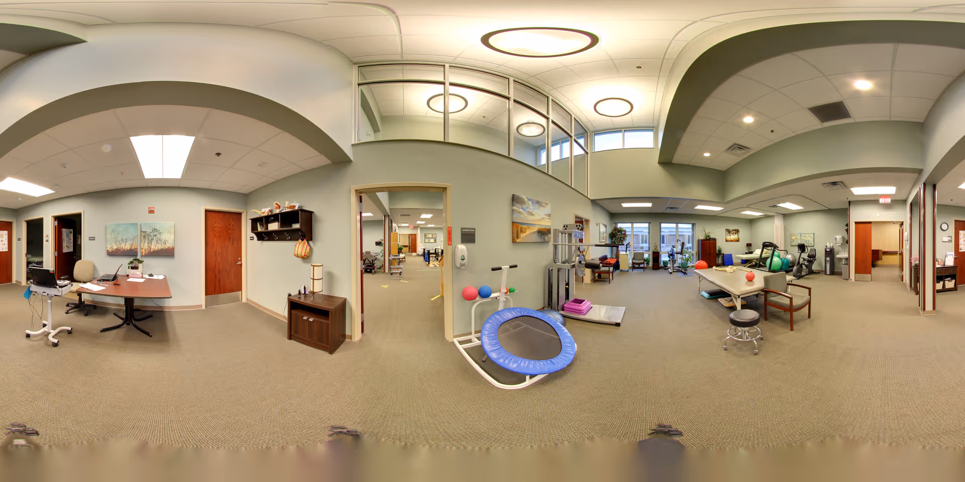 Interior of a senior living facility rehabilitation/exercise room with therapy equipment, treatment tables, and desks.