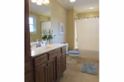 A clean bathroom with a wooden vanity featuring a white countertop and sink, a mirror above the sink, a toilet with a blue seat cover, a blue bath mat on the floor, and a shower with a white curtain. There is a small window and a framed picture on the wall.
