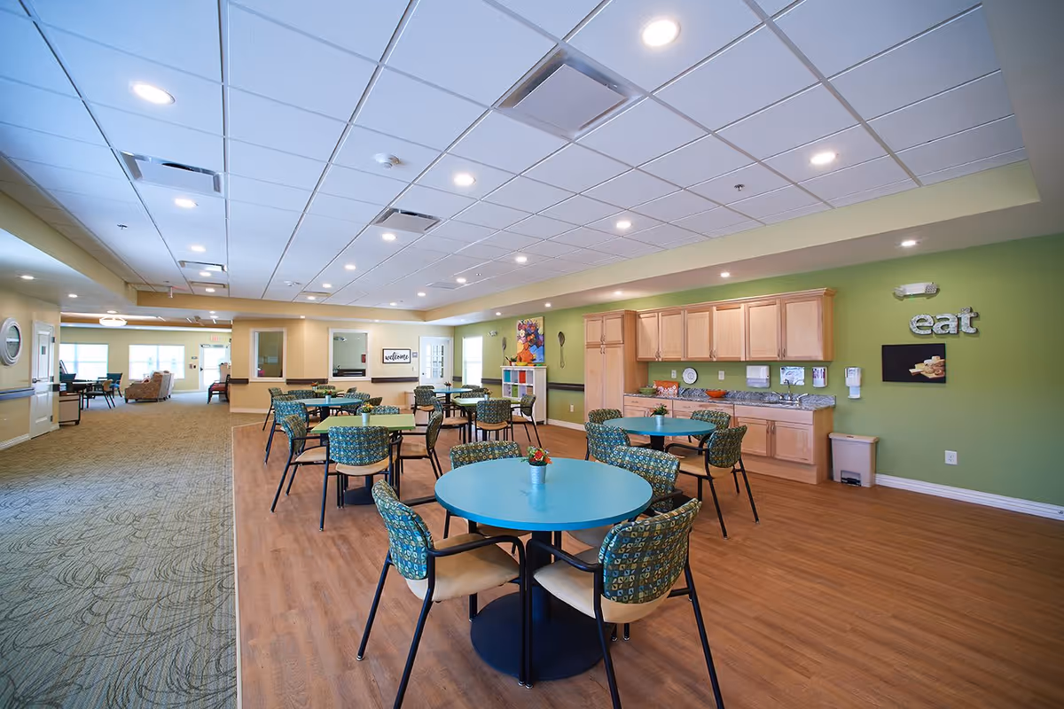 Bright communal dining area with round tables and patterned chairs and a kitchenette against a green wall with an "eat" sign.