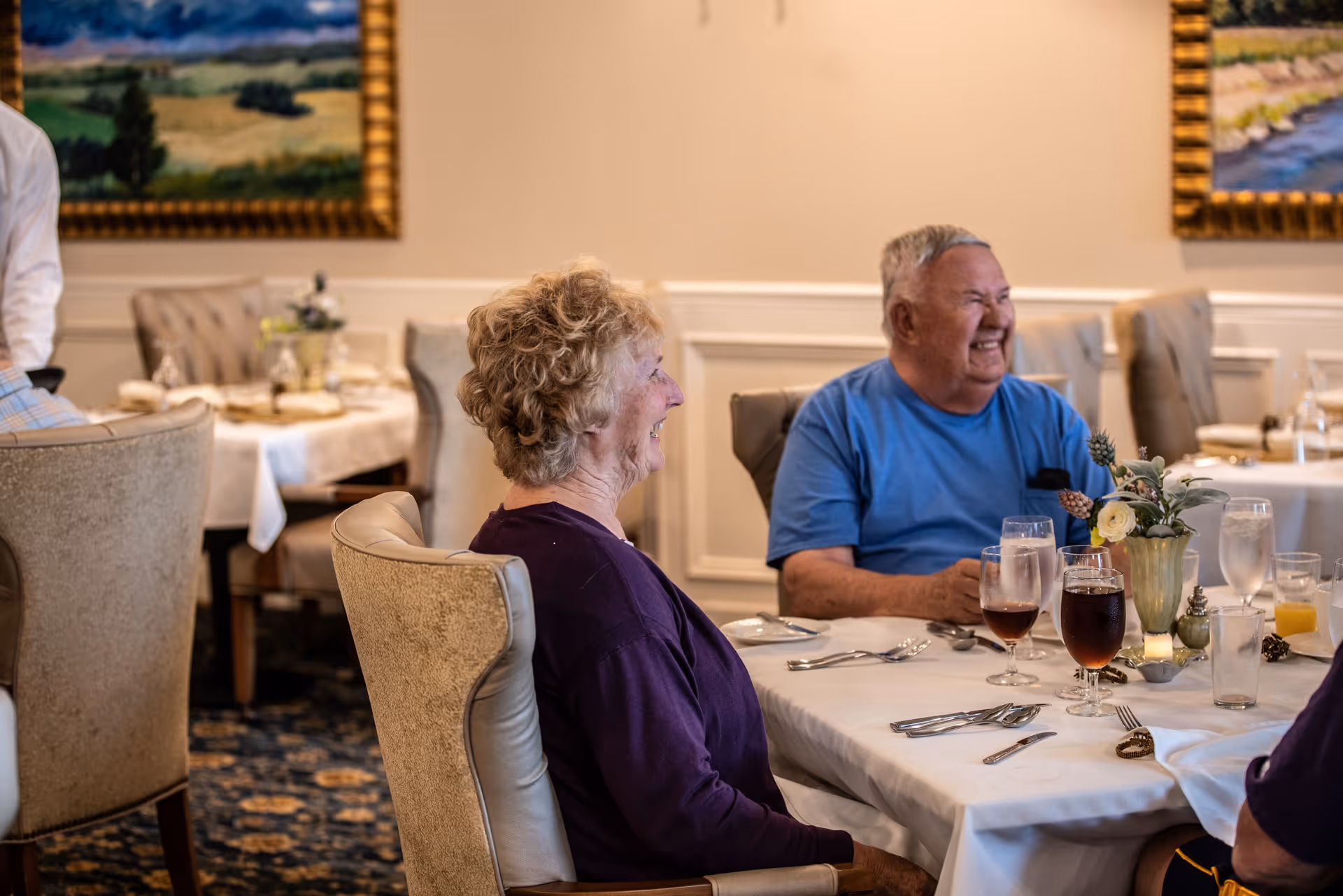 Two elderly people sitting at a dining table in a well-decorated dining room, smiling and enjoying their time. The table is set with glasses of iced tea, water, and silverware, with a floral centerpiece. The background shows framed landscape paintings and upholstered chairs.