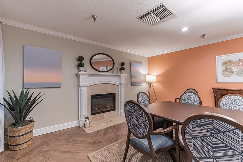 A cozy dining area with a rectangular wooden table surrounded by six patterned chairs. The room features a white fireplace with a mirror and decorative plants on the mantel, two framed pictures on the walls, a floor lamp in the corner, and a large potted plant near the window. The walls are painted in beige and orange tones, and the floor has a wood finish with a woven rug under the table.