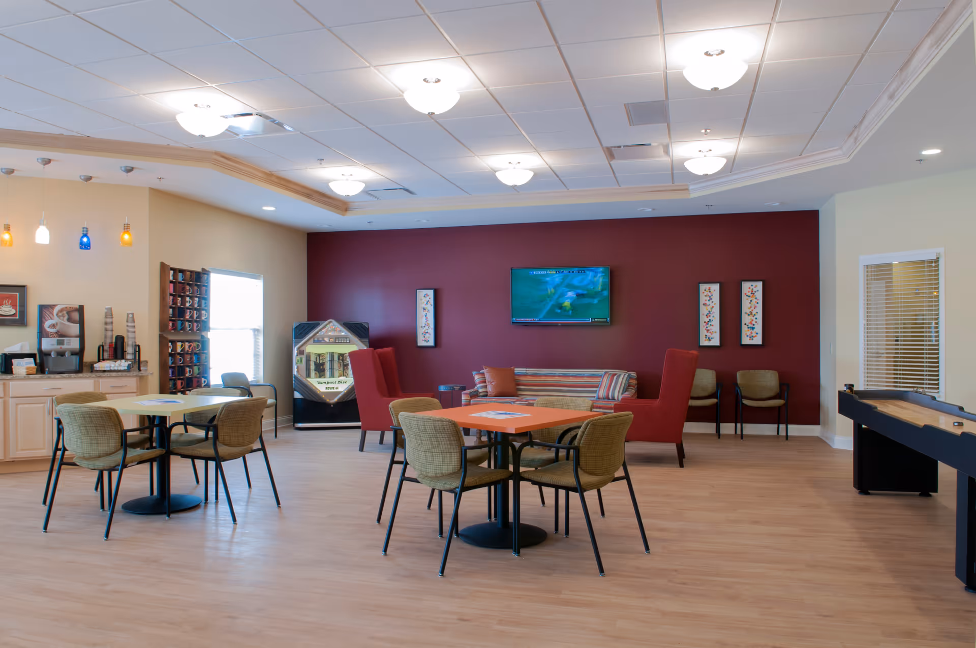 A spacious common area with light wood flooring, two tables each surrounded by four chairs, a coffee station with colorful mugs on shelves, a jukebox, a striped sofa with two red armchairs, a wall-mounted TV, and a shuffleboard table on the right side.