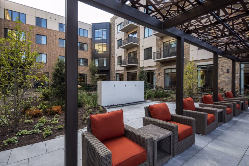 Outdoor seating area at Eagleview Landing featuring a row of wicker chairs with red cushions under a pergola, surrounded by landscaped gardens and a multi-story building with balconies and large windows in the background.