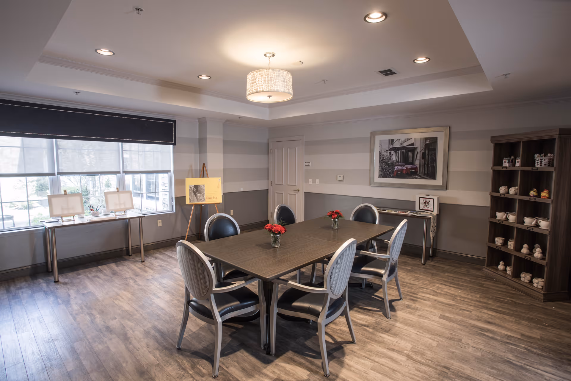 Communal dining/activity room with a rectangular table surrounded by chairs, small vases of red flowers, shelving with ceramics, and large windows.